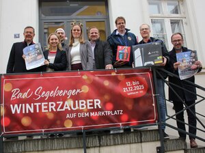 Eine Gruppe von Menschen steht auf einer Treppe vor einem Gebude mit Glastr. Sie halten Plakate und Kartons mit Spielkonsolen. Vor ihnen ist ein rotes Banner mit weier Schrift befestigt, auf dem steht: 'Bad Segeberger Winterzauber auf dem Marktplatz' sowie '12. bis 21.12.2025'.