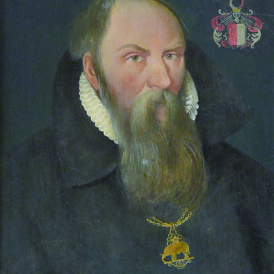 Bild vergr&ouml;&szlig;ern: Portr�t eines Mannes mit langem Bart, schwarzem Mantel, wei�em R�schenkragen und goldener Kette mit Anh�nger, daneben ein Wappen mit roten und schwarzen Elementen.