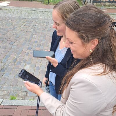 Bild vergr&ouml;&szlig;ern: Zwei Frauen stehen auf einem gepflasterten Gehweg und schauen auf ihre Smartphones.