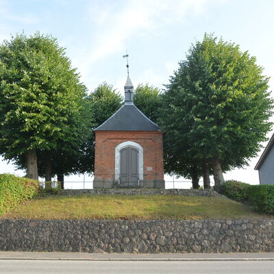 Bild vergr&ouml;&szlig;ern: Kleine Backsteinkapelle mit grauem T�rbogen und Dachreiter, umgeben von gro�en B�umen auf einem erh�hten, mit Steinmauer eingefassten Grundst�ck zwischen Wohnh�usern.