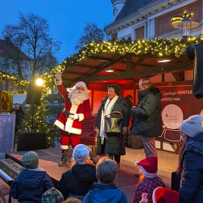 Bild vergr&ouml;&szlig;ern: Weihnachtsmann auf einer mit Lichterketten geschm&uuml;ckten B&uuml;hne beim Weihnachtsmarkt Bad Segeberger Winterzauber, umgeben von zwei Personen und einem Publikum mit Kindern in Winterkleidung und Weihnachtsm&uuml;tzen.
