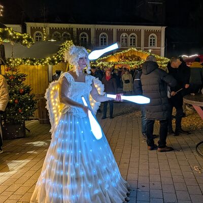 Bild vergr&ouml;&szlig;ern: Eine Person in einem wei&szlig;en, mit Lichtern geschm&uuml;ckten langen Kleid jongliert leuchtende Keulen auf dem Weihnachtsmarkt Bad Segeberger Winterzauber, im Hintergrund sind festlich beleuchtete St&auml;nde und Besucher zu sehen.