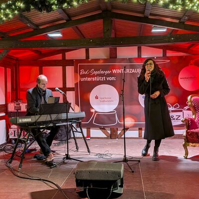 Bild vergr&ouml;&szlig;ern: Eine B&uuml;hne mit Weihnachtsbeleuchtung, auf der ein Musiker am Keyboard und eine S&auml;ngerin mit Mikrofon auftreten, im Hintergrund ein Banner mit der Aufschrift 'Bad Segeberger Winterzauber unterst&uuml;tzt von'.