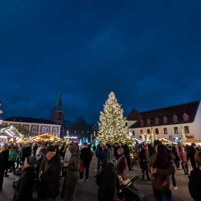 Bild vergr&ouml;&szlig;ern: Weihnachtsmarkt mit beleuchtetem Weihnachtsbaum in der Mitte, umgeben von St&auml;nden und zahlreichen Besuchern unter dunklem Abendhimmel in Bad Segeberg.