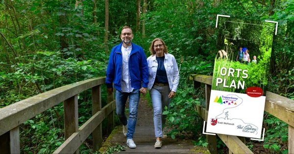 Bild vergr&ouml;&szlig;ern: Zwei Personen gehen auf einem Holzsteg durch einen Wald, rechts steht ein Schild mit der Aufschrift 'ORTS PLAN'.