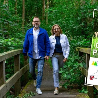 Zwei Personen gehen auf einem Holzsteg durch einen Wald, rechts steht ein Schild mit der Aufschrift 'ORTS PLAN'.