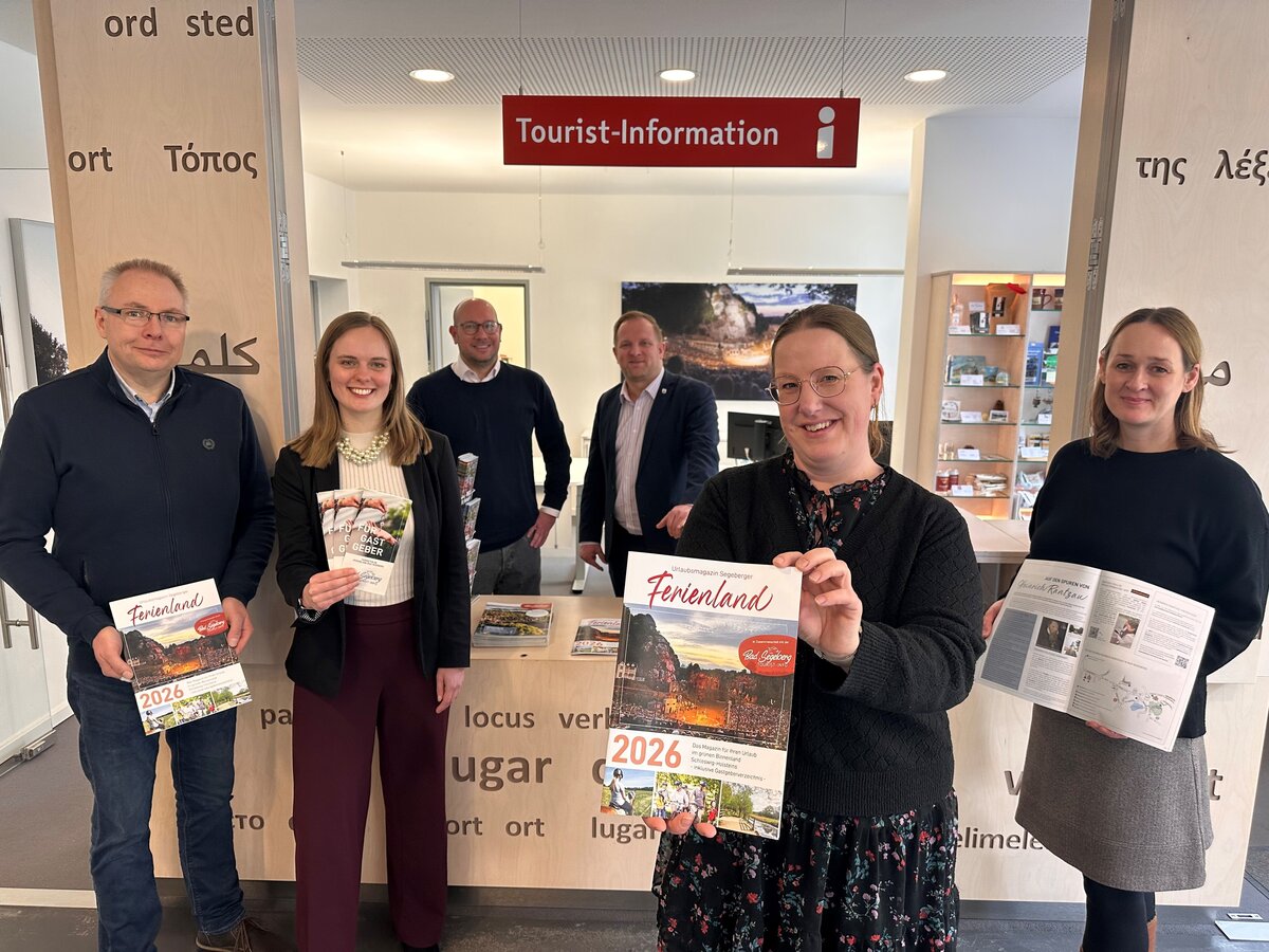 Sechs Personen in einem Tourist-Informationsb&uuml;ro halten Urlaubsmagazine mit dem Titel 'Ferienland 2026' in der Hand, im Hintergrund ein rotes Schild mit der Aufschrift 'Tourist-Information'