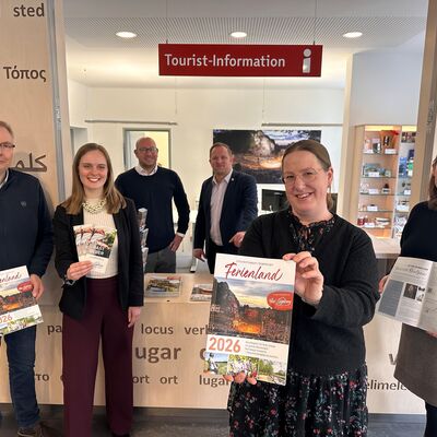 Sechs Personen in einem Tourist-Informationsb&uuml;ro halten Urlaubsmagazine mit dem Titel 'Ferienland 2026' in der Hand, im Hintergrund ein rotes Schild mit der Aufschrift 'Tourist-Information'